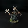 A miniature figure of a bearded dwarf-like character with long, dark hair and a thick beard, wearing detailed armor and holding two large, ornate hammers with intricate designs. The figure stands on a circular base with small patches of grass and textured ground. The background is black.