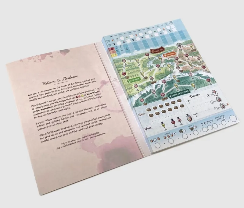 Open book with a light pink left page featuring black text under the heading "Welcome to Bordeaux" and a white right page displaying a colorful illustrated map of Bordeaux vineyards, wine glasses, barrels, and tokens. The map includes labeled areas such as "Left Bank" and "Entre-Deux-Mers," along with icons for resources like grapes and casks. At the top of the right page, there is a grid with letters and numbers for tracking. The bottom of the left page has instructions to flip to the inside cover for solo rules and campaigns.