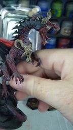 A detailed dragon miniature is being held in a hand, showcasing its intricate features, including prominent scales, fierce facial expression, and large wings with a red underside. The dragon is painted in dark hues with highlights that accentuate its muscular form and menacing appearance. In the background, various paint jars are visible, indicating a workspace for model painting.