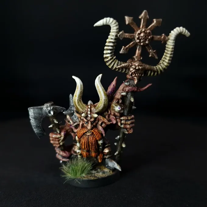 A miniature figure of a Chaos Dwarf with a long orange beard, wearing armor and holding a large axe in one hand and a staff topped with a ram's skull and circular blade in the other. The figure stands on a small base with green tufts of grass. The background is black.