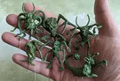 A hand holds several green resin miniatures, showcasing a variety of fantastical creatures. The figures include grotesque designs with multiple limbs, horns, and exaggerated features, reflecting an old-school aesthetic reminiscent of classic tabletop miniatures. Each miniature is mounted on a small pin for display, emphasizing their intricate details and unique forms.