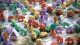 Colorful plastic yak miniatures in various poses on a hexagonal board with light blue and white terrain. Some yaks wear decorative headpieces in gold, purple, and red. Small brown and green tokens with floral designs are scattered among them.