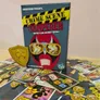 Crime Scene Tamperer game box with cards and tokens scattered on a wooden floor.