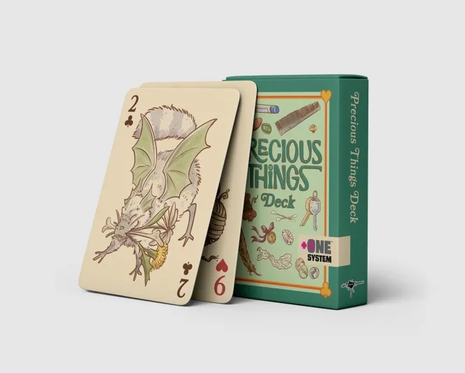 A deck of playing cards from the game "Precious Things" is displayed alongside its box. The box is green with whimsical illustrations of mundane objects like buttons and paperclips, and features the title "Precious Things Deck" along with the "+One System" logo. Two cards are shown prominently: one depicts a small dragon holding a flower, while the other shows a number and suit, indicating their use in gameplay.