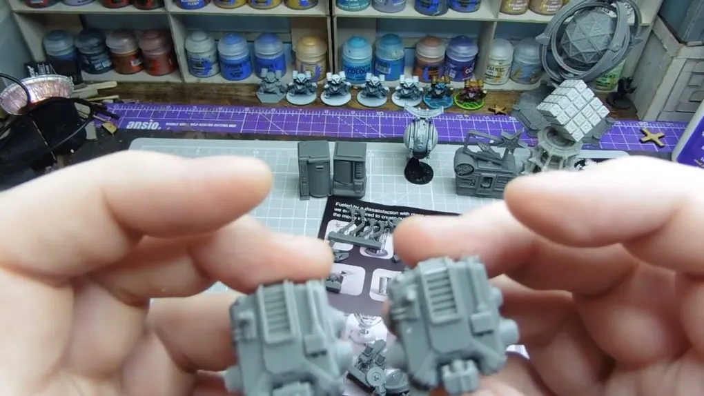 Two gray miniature components are held in each hand, showcasing intricate details such as ridges and small protrusions. In the background, various other gray miniatures and model parts are arranged on a crafting mat, with a selection of paint bottles visible on shelves above. A printed sheet with diagrams and instructions is partially visible beneath the components.