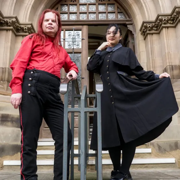 Two individuals pose in front of a grand, gothic-style building. One person, wearing a bright red shirt and black trousers with red stripes, stands confidently with arms crossed. The other person, dressed in a long black coat with a high collar and a blue shirt underneath, holds the hem of their coat, creating a dramatic silhouette. The ornate entrance of the building, featuring large wooden doors and intricate stonework, serves as a striking backdrop.