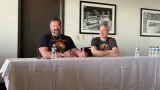 Two men are seated at a table during a panel discussion. On the left, a man with a beard, wearing a black shirt featuring a colorful graphic and a necklace, gestures with his hands while speaking. On the right, a man with short gray hair, dressed in a gray t-shirt with a retro design, smiles and listens attentively. Behind them, a framed black-and-white photograph of a decorative railing is visible on the wall. A bottle of water and a small device are placed on the table in front of them.