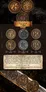 Three antique-style metal coins, each 30 mm in diameter, displayed front and back. Fronts show intricate designs with figures and text. Backs appear tarnished. Coins mounted on a dark, textured surface with a sword handle.