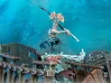 A dynamic scene features an Idoneth Deepkin warrior, the Ikon of the Sea, poised on a wave of water, wielding a curved sword and adorned with intricate armor and a decorative headdress. In the background, remnants of a shipwreck are visible, with splashes of water and a menacing sea creature emerging from the waves. The overall color palette includes vibrant blues and earthy tones, enhancing the aquatic theme of the model.