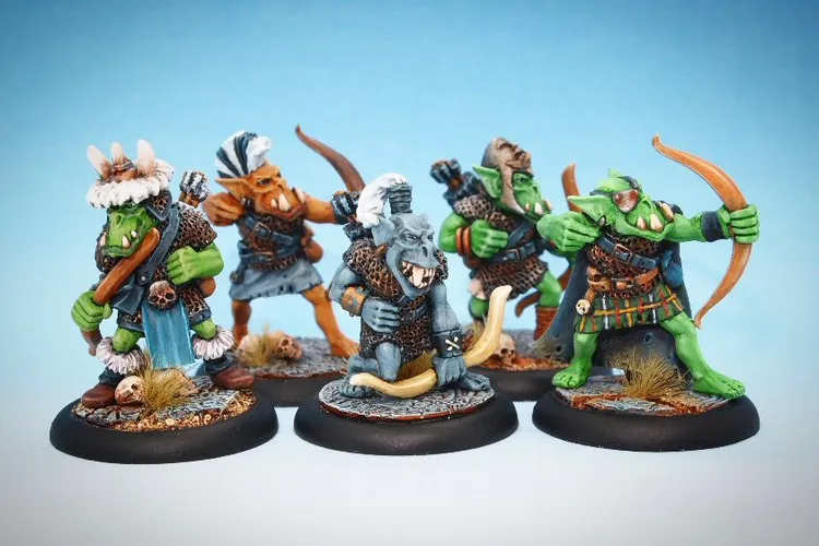 Five colorful orc miniatures are displayed against a light blue background. Each orc is uniquely posed and armed, with one holding a bow, another wielding a club, and others equipped with various weapons like spears and crossbows. They are dressed in a mix of armor and clothing, featuring details like fur trim and decorative elements, set on textured bases with grass and skull accents.