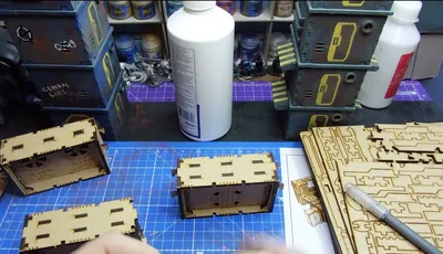 A workbench scene features a partially assembled laser-cut building structure made of light wood, positioned in the foreground. Behind it, a stack of completed structures in various colors, including blue and yellow, is visible, alongside bottles of paint and glue. A cutting tool rests on the table, and a printed reference image of a similar structure is partially visible.