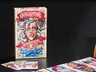 The image features the box of the card game "Guillotine," prominently displaying an illustration of a startled woman with gray hair and a blue bow, set against a light-colored background. Next to the box, a wooden guillotine prop is shown, designed to hold the noble cards. In the foreground, several tarot-sized noble cards are laid out, showcasing colorful illustrations of various characters, each with distinct expressions and attire, reflecting the game's theme of the French Revolution.