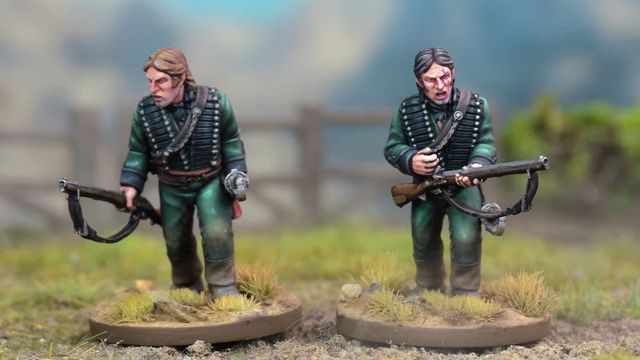 Two detailed miniatures of soldiers from the Sharpe’s Rifles series are displayed on textured bases, set against a blurred natural background. Both figures wear green uniforms with black crossbelts; one holds a rifle and has a serious expression, while the other appears to be in a more contemplative pose. The bases feature tufts of grass, enhancing the realistic terrain effect.