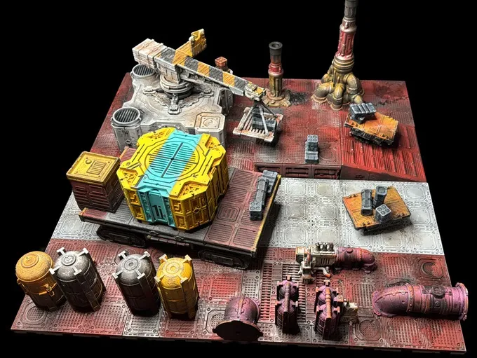 A detailed modular sci-fi terrain setup features various structures and elements, including a large yellow and blue container, a crane, and several industrial tanks in shades of gray and yellow. The terrain is painted with a mix of red and gray tones, creating a weathered, industrial look. Additional components like pipes and small vehicles are scattered throughout, enhancing the scene's complexity and depth for tabletop gaming.