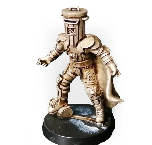 A gold-colored miniature figurine of a character wearing a cylindrical helmet resembling a trash bin, with a cape flowing behind. The figure stands on a round black base with a textured surface. The helmet has a handle on top and a small emblem on the front. The figure is posed with one arm extended forward and one leg slightly bent, as if mid-step.