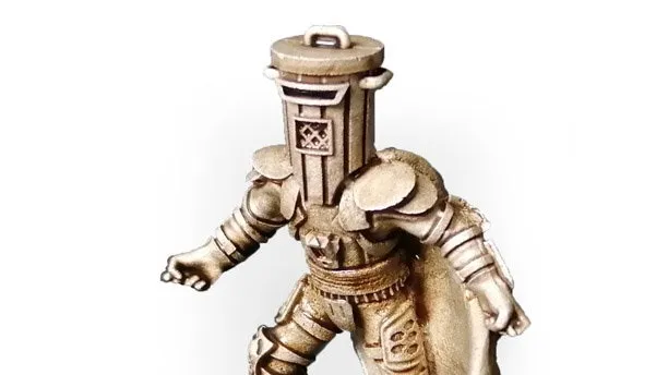 A gold-colored miniature figurine of a character wearing a cylindrical helmet resembling a trash bin, with a cape flowing behind. The figure stands on a round black base with a textured surface. The helmet has a handle on top and a small emblem on the front. The figure is posed with one arm extended forward and one leg slightly bent, as if mid-step.