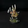 A painted miniature of a Chaos Dwarf with a long yellow beard, wearing metallic armor and a horned helmet. The figure holds a staff with a twisted design and stands on a circular base with small patches of grass and rocks. The background is black.