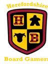 A shield-shaped logo features a divided design with four quadrants. The top left quadrant displays a red die with black dots, the top right features a black meeple, the bottom left shows a black cow silhouette, and the bottom right has a red die with white dots. The text "Herefordshire Board Gamers" is prominently displayed above and below the shield in a bold, yellow font.