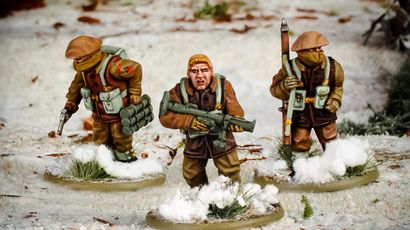 Three multi-part British and Canadian winter infantry figures are depicted in a snowy landscape. The central figure, with short blond hair, is holding a rifle and wearing a brown coat, while the other two figures are dressed in winter gear, one carrying a set of explosives and the other holding a sidearm. The bases of the figures are adorned with patches of snow and tufts of grass, enhancing the wintery scene.