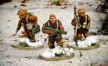 Three multi-part British and Canadian winter infantry figures are depicted in a snowy landscape. The central figure, with short blond hair, is holding a rifle and wearing a brown coat, while the other two figures are dressed in winter gear, one carrying a set of explosives and the other holding a sidearm. The bases of the figures are adorned with patches of snow and tufts of grass, enhancing the wintery scene.