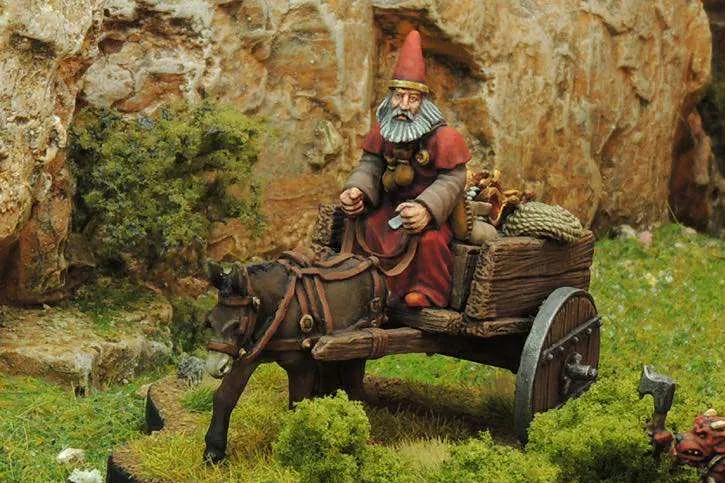 A miniature figure of a gnome with a long beard and a red pointed hat sits atop a wooden cart pulled by a dark brown donkey. The cart is laden with various items, including sacks and a small barrel, and is set against a backdrop of rocky terrain and green foliage. The scene captures a whimsical, fantasy-inspired atmosphere, perfect for a tabletop RPG setting.