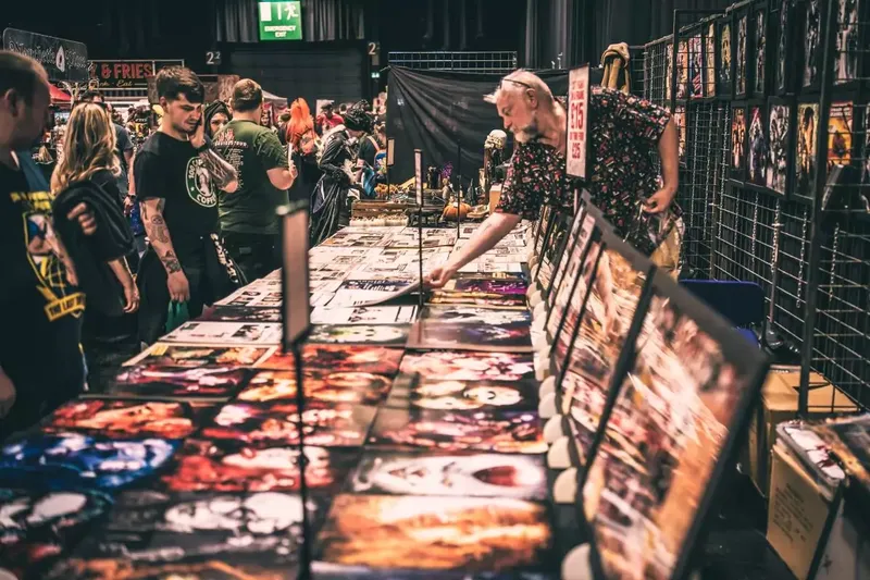 A bustling convention scene filled with attendees exploring various stalls and displays. In the foreground, a long table is covered with vibrant artwork and prints, showcasing horror-themed images, while a person examines the items closely. The background features a crowd of people, some in themed attire, engaging with the immersive atmosphere of the event.