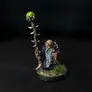 A miniature figure of a bearded dwarf wearing a dark robe, holding a staff topped with a glowing green orb. The staff is detailed with a twisted, thorn-like design. The figure has goggles over its eyes and stands on a small circular base with patches of green grass. The background is black.