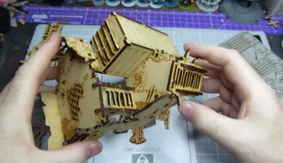 A partially assembled Orc Lookout tower made of laser-cut wood is being held by two hands. The structure features multiple levels, with a slanted roof and wooden slats, showcasing intricate details like windows and a skull motif. In the background, a crafting mat and various miniatures are visible, indicating a tabletop gaming environment.