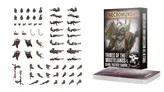 A collection of miniature parts and accessories for the Necromunda game is displayed, focusing on the Ash Wastes nomads. On the left, various weapon components and heads are arranged, showcasing a range of designs including rifles and melee weapons. On the right, the "Tribes of the Wasteland" book and gang tactics cards are prominently featured, with artwork depicting a nomad character.