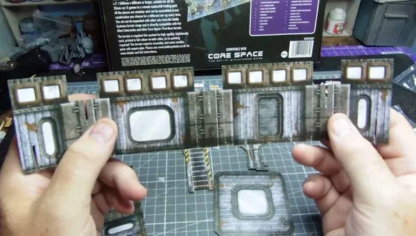 A hand holds a piece of unpainted cardboard terrain with a rectangular window and weathered metal texture. On a green cutting mat, additional assembled cardboard terrain pieces with similar windows, ladders, and industrial details are visible. In the background, a product box for "CORE SPACE" by Battle Systems is partially visible, with text and a barcode.