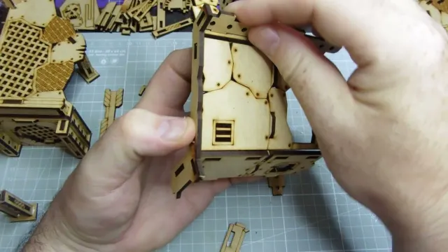 A hand holds a piece of laser-cut wood, part of the TT Combat Orc Mega Bunker model, showcasing intricate details like a small window and textured surfaces. Surrounding the hand are various other wooden components and parts of the model, laid out on a cutting mat with grid lines. The overall scene emphasizes the assembly process of the Orc Sektor level 2 build.
