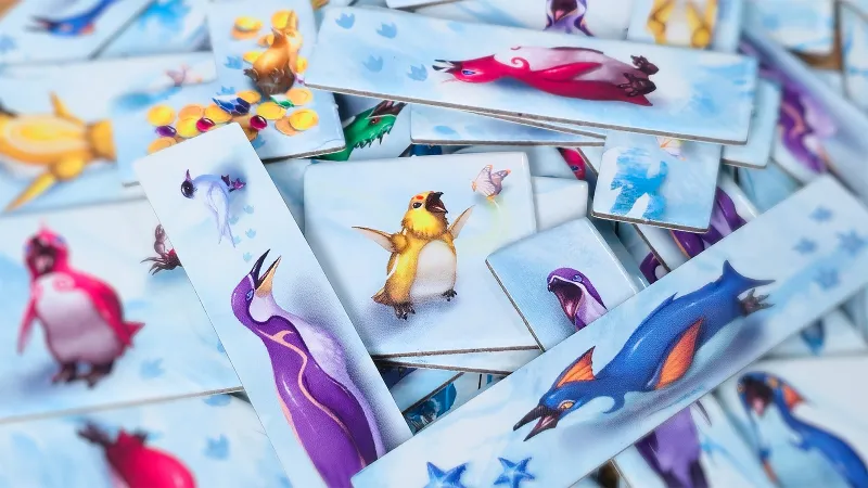 A scattered arrangement of rectangular game tiles featuring colorful, cartoon-style illustrations of various penguins and other creatures. The central tile shows a yellow-and-white penguin with outstretched wings, surrounded by tiles depicting a purple-and-blue penguin, a green dragon, a red-and-pink sea creature, and other vibrant animals. Some tiles include small decorative elements like butterflies or coins. The background is a light blue with subtle cloud-like patterns.