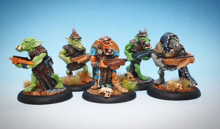 A group of five intricately painted orc miniatures stands against a light blue background. The orcs display a variety of features, including different armor styles and weapons, such as crossbows and hand weapons. The bases are adorned with grass and small skulls, adding to the overall detail and character of each figure.