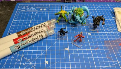 Three Playmarkers from AK are displayed on a blue cutting mat, featuring a grid pattern. The markers are in red, blue, and white, each labeled "PLAYMARKERS FOR TABLETOP GAMERS." In the foreground, two small miniatures are positioned on clear bases: a black creature and a red creature, while a larger green creature and a blue figure are visible in the background.