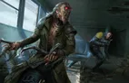 A monstrous humanoid figure with glowing red eyes, exposed muscle, and jagged teeth, wearing a dark green trench coat, holding a large weapon. Behind it, a smaller armored figure with a helmet and yellow emblem kneels, aiming a weapon. Dark, cluttered indoor setting with dim lighting and debris.