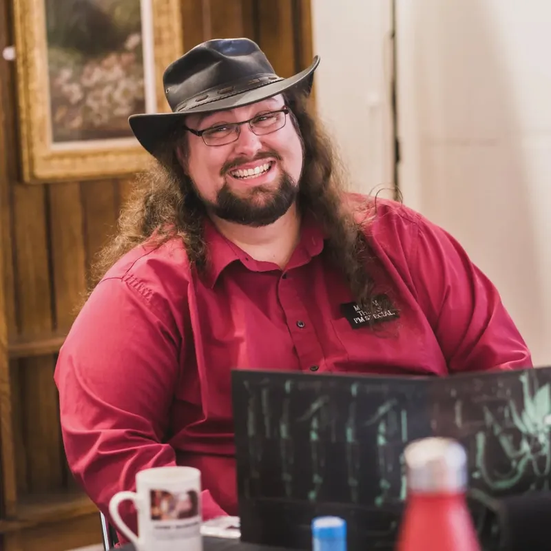 A smiling individual with long, curly hair and a beard is seated at a table, wearing a black hat and a red shirt. They are positioned in front of a dark screen displaying game elements, with a mug and various bottles visible on the table. The background features wooden paneling and a framed artwork, contributing to a cozy, inviting atmosphere.