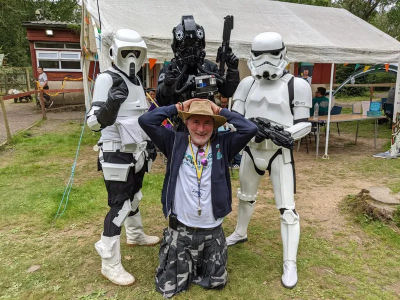 A man in a wide-brimmed hat and a casual outfit kneels in the foreground, smiling and playfully raising his hands above his head. Behind him, two Stormtroopers and a TIE Fighter pilot in full costume stand with their weapons, creating a fun and engaging atmosphere. The setting features a grassy area with a tent in the background, suggesting a lively outdoor event.