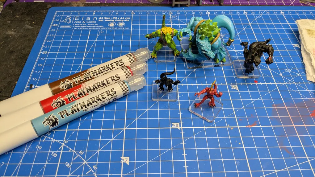 Three Playmarkers from AK are displayed on a blue cutting mat, featuring a grid pattern. The markers are in red, blue, and white, each labeled "PLAYMARKERS FOR TABLETOP GAMERS." In the foreground, two small miniatures are positioned on clear bases: a black creature and a red creature, while a larger green creature and a blue figure are visible in the background.