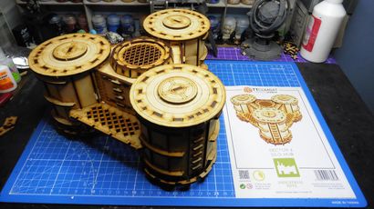 A detailed model of the Sector 3 Silo Hub from TT Combat is prominently displayed on a blue cutting mat. The structure features multiple cylindrical silos with intricate wooden textures and circular tops, showcasing a combination of open latticework and solid panels. In the background, various paint pots and a few other miniature models are visible, along with the instruction sheet for the Silo Hub, which includes an illustration of the assembled model.