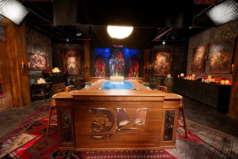 A richly designed tabletop setting features a large, intricately carved wooden table at the center, adorned with a blue surface that suggests a gaming area. Surrounding the table are various chairs, and the backdrop includes stone walls with colorful stained glass windows, casting vibrant light across the scene. The space is decorated with candles, artwork depicting fantasy themes, and a patterned rug, creating an immersive atmosphere suitable for a Dungeons & Dragons actual play show.