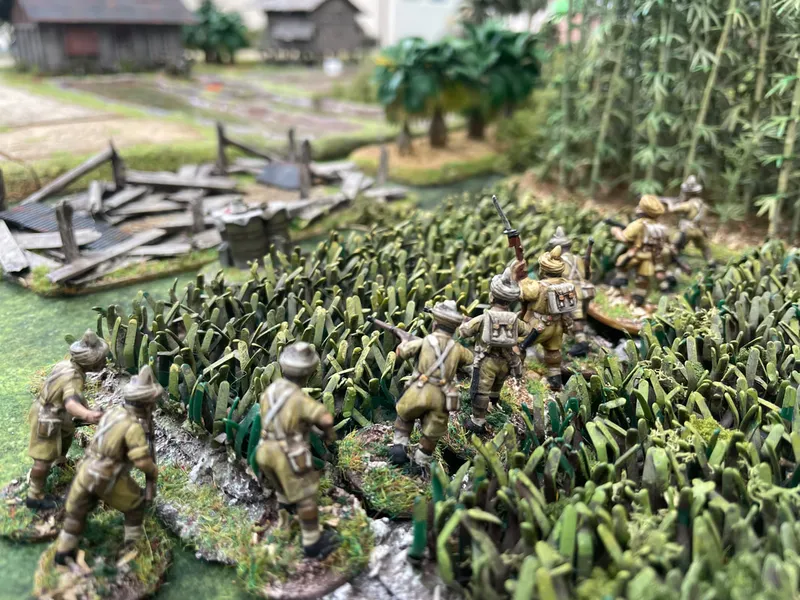 A group of miniature soldiers in khaki uniforms is advancing through dense green foliage, with some figures holding rifles and others appearing to be in various poses of movement. The background features a detailed landscape with palm trees and a rustic building, enhancing the immersive setting of the wargaming scene. The terrain is richly textured, with a mix of grass and underbrush, creating a realistic battlefield environment.
