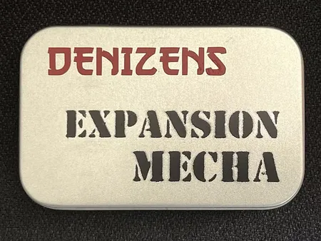 A small, silver tin features the text "DENIZENS" in bold red letters at the top and "EXPANSION MECHA" in large, black, stencil-style font below. The tin's surface has a smooth, metallic finish, and it is set against a dark background, emphasizing its compact and portable design. This packaging represents the new expansion for the Space Denizens wargame, which includes mechs and terrain features.