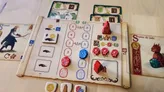 A board game setup for Greylune showing a player’s board with numbered action slots (1–5), icons for lightning, coins, and a blue "U" symbol. Wooden tokens include a red pyramid, red and blue circular pieces, and small house and rose-shaped markers. Two card displays show "0" and "2" victory points.
