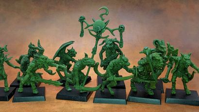 A collection of old school miniatures from Satyr Art Studio, featuring various fantastical creatures in a vibrant green color. The figures include beastmen with distinct features such as horns and claws, some wielding weapons, and a central figure holding a staff topped with a skull and a crescent. They are displayed on black bases against a warm, textured background.