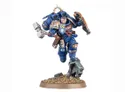 A dynamic miniature of a Space Marine captain in blue armor, poised mid-action with a thunder hammer in one hand and a relic shield in the other. He features a jump pack on his back and has a determined expression, with details like a skull emblem on his chest and a decorative shoulder pad. The base is textured with dirt and small rocks, adding to the overall dramatic presentation.