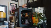 A modular, 3D-printable display case, designed for collectors, is shown in the image. The case features a sleek black design with a snap-fit assembly, and is currently being handled by a person, showcasing its ease of use. Inside the case, various collectibles are displayed, including a Charizard figure, a camera, and trading cards, while decorative items and framed artwork are visible in the background.
