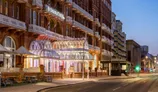 A grand hotel entrance features an ornate glass canopy with decorative lighting, flanked by brickwork and balconies of the building. The DoubleTree Metropole Hotel is prominently displayed above the entrance, while flower pots add a touch of color to the scene. The street is quiet, illuminated by soft evening lights, creating a serene atmosphere as dusk settles.