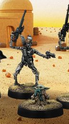 A group of four miniatures from the Star Wars universe is displayed against a desert backdrop featuring a simple structure. On the left, a character in a silver and black armor, resembling a bounty hunter, stands confidently. Next to him, two IG-Series Assassin Droids are posed with weapons drawn, showcasing their metallic bodies and intricate details. In the foreground, Grogu, a small figure with large ears, is depicted holding a small object, adding a whimsical touch to the scene. The ground is textured with grass and dirt, enhancing the overall aesthetic of the miniatures.