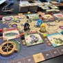 A board game setup with a map featuring numbered hexagonal tiles (83, 84, 85, 86, 87). Miniature figures, including a horse and a person, are placed on the board. Various illustrated cards depict plants, animals, and locations, with text including "Tithymalus" and "Carl von Linné."