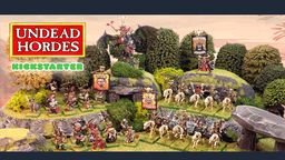 The scene features a vibrant tabletop display showcasing the "Undead Hordes" miniatures from Red Bard Games' Kickstarter campaign. In the foreground, a variety of skeletons and zombies are arranged on grassy terrain, with some holding banners and others in dynamic poses. A prominent liche king rides a zombie dragon atop a rocky outcrop, while additional figures, including a necromancer and various undead warriors, populate the scene, surrounded by lush greenery and stone formations. The overall composition exudes a classic fantasy aesthetic, emphasizing the theme of an undead army ready for battle.